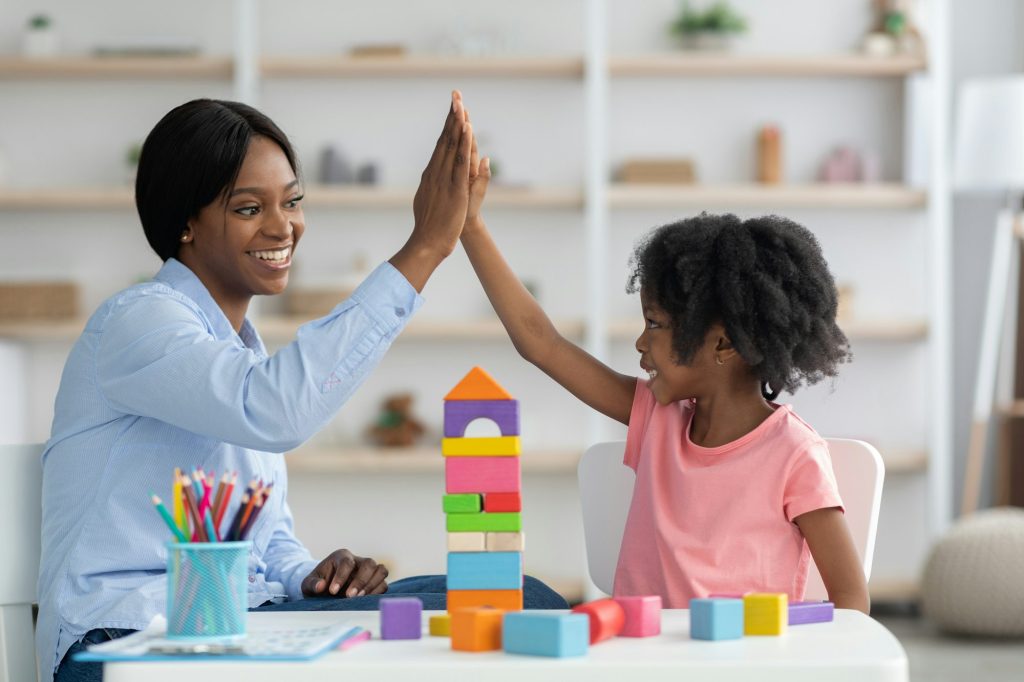 Pretty preschool teacher exercising with happy little girl at kindergarten